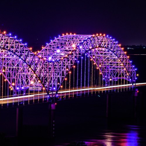 A brightly lit bridge with colorful lights arching over a river at night, reflecting on the water below.