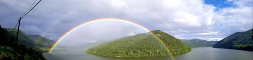 A vibrant double rainbow arcs over a lush green island in a serene lake, surrounded by mountains and cloudy skies.
