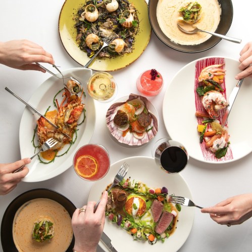 A table spread with various gourmet dishes and drinks, hands reaching for food with forks.