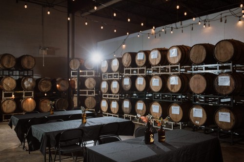 A spacious venue with wooden barrels stacked in the background and tables set with flowers in front, illuminated by string lights.