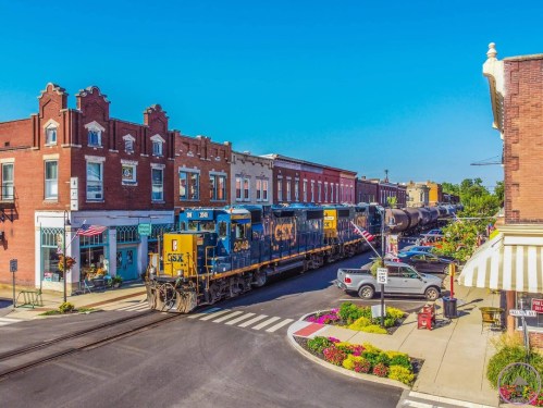 A CSX train passes through a charming town with colorful storefronts and flower beds under a clear blue sky.