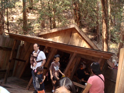 A tilted wooden structure in a forest, with people walking around, including a man carrying a baby and children exploring.