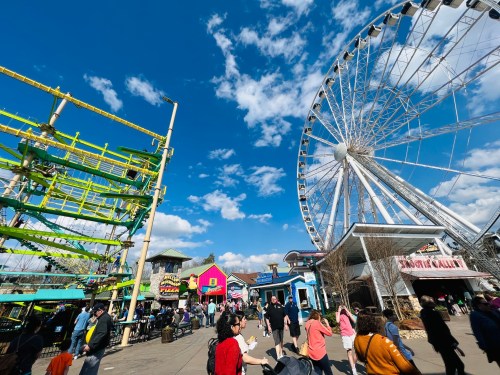 A bustling amusement park scene featuring a large Ferris wheel, colorful buildings, and crowds of people enjoying the day.