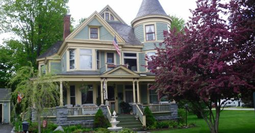 Victorian-style house with a turret, surrounded by trees and a fountain, featuring an American flag on the porch.