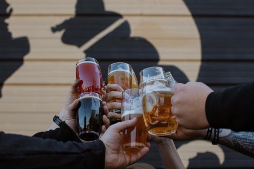 A group of hands holding various glasses of beer in a toast against a mural backdrop.