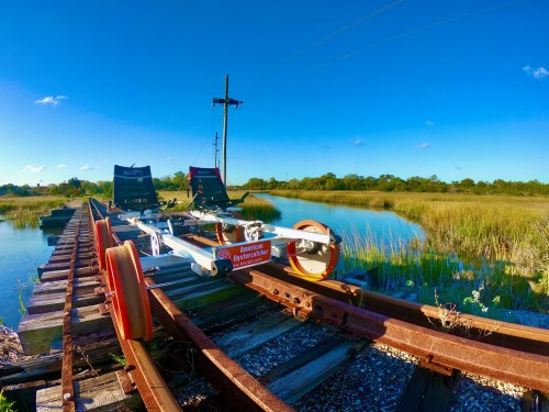 A view of a wooden dock with a rail cart on tracks, surrounded by water and tall grass under a clear blue sky.