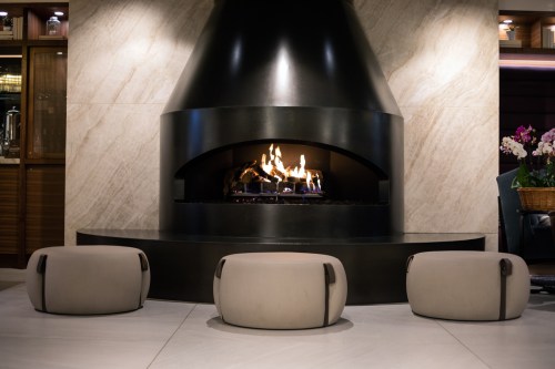 A modern fireplace with a sleek design, surrounded by three round, neutral-colored stools. Warm flames flicker inside.