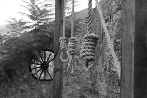 Black and white image of three coiled ropes hanging near a stone wall and a wooden wheel.
