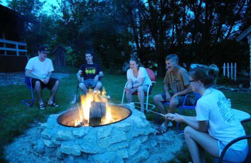 A group of five people sitting around a campfire, roasting marshmallows in a serene outdoor setting at dusk.