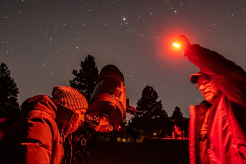 A person looks through a telescope while another holds a red light, set against a starry night sky.