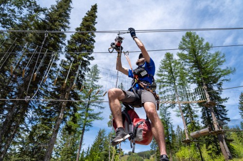 A person in safety gear climbs a rope course among tall trees under a blue sky.