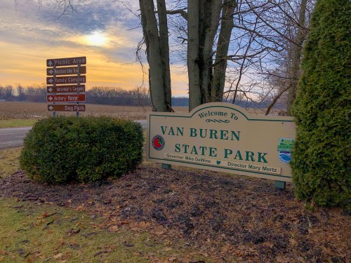 Welcome sign for Van Buren State Park, with directional signs and a scenic background of trees and a sunset.