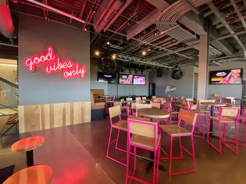 Interior of a lively bar with pink chairs, a "good vibes only" neon sign, and screens displaying entertainment.