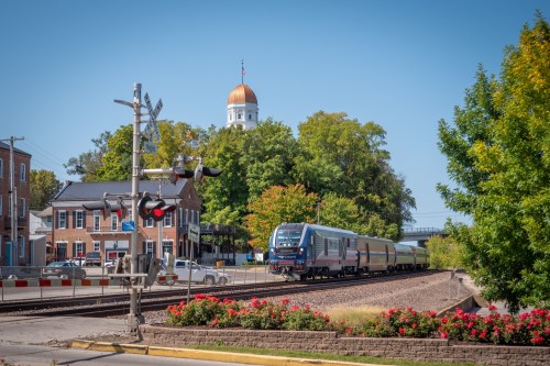 A train approaches a crossing near a small town, with a golden dome building and trees in the background.