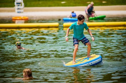 A boy balances on a paddleboard in a lake, while others swim and kayak in the background on a sunny day.