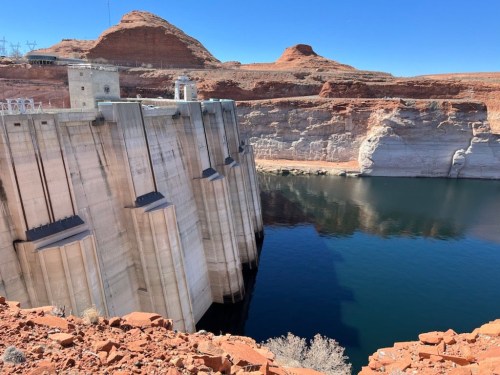 A large concrete dam stands beside a calm blue lake, surrounded by red rock formations under a clear blue sky.