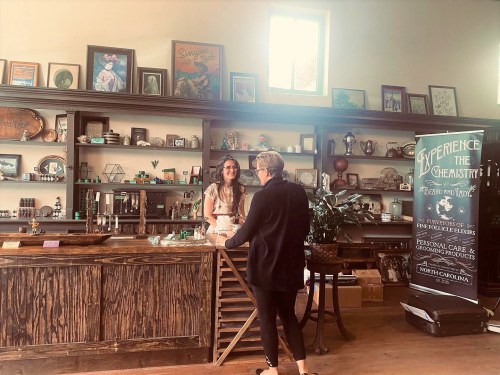 A woman in a white dress talks to another woman at a wooden counter in a shop filled with various items and decor.