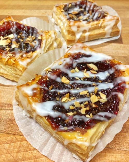 Three flaky pastries topped with jam, drizzled with icing, and sprinkled with chopped nuts, resting on paper liners.
