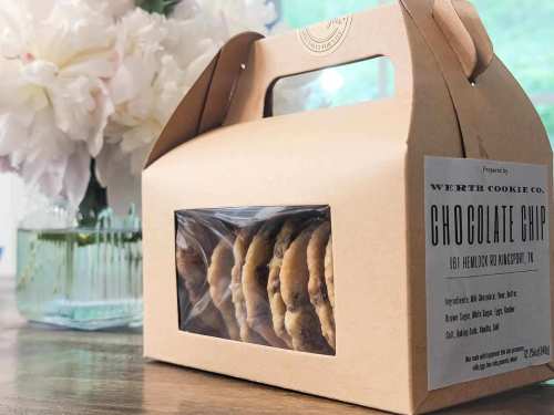 A box of chocolate chip cookies with a clear window, placed on a table near flowers.