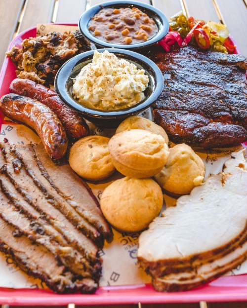 A vibrant platter of barbecue featuring brisket, sausage, pulled pork, baked beans, coleslaw, and cornbread muffins.