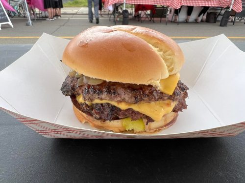 A large cheeseburger with two beef patties, melted cheese, pickles, and onions, served in a paper tray.