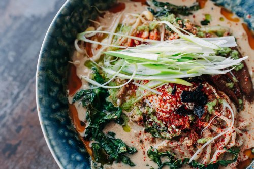 A bowl of noodles topped with greens, scallions, and spices in a creamy sauce, set on a wooden table.