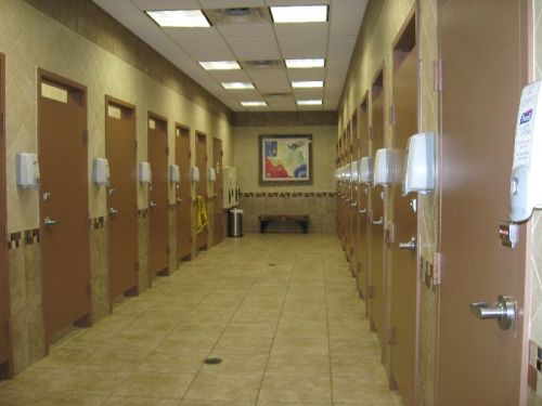 A hallway with multiple restroom doors, hand dryers, and tiled floors, featuring a painting on the wall.