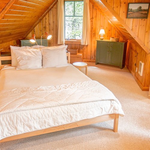 Cozy attic bedroom with wooden walls, a bed, nightstand, and soft lighting from lamps and a window.