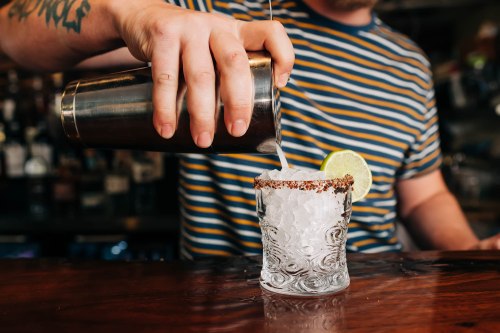 A bartender pours a cocktail into a glass with a salted rim, garnished with a lime slice.