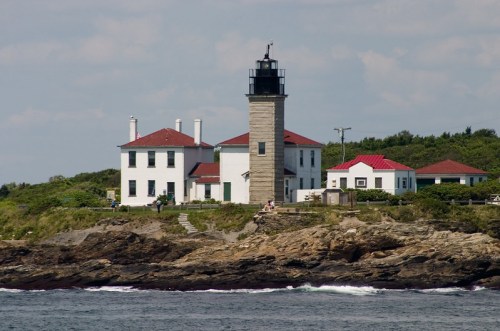 A lighthouse on a rocky shore, surrounded by greenery and buildings with red roofs under a blue sky.