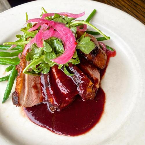 A plate of sliced duck breast topped with pickled onions, served on a bed of green beans and drizzled with berry sauce.
