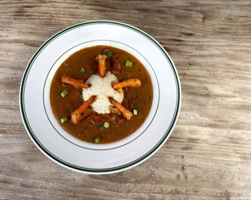 A bowl of brown soup with shrimp and a mound of white rice, garnished with green onions, on a wooden surface.