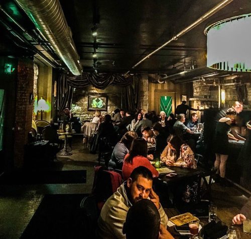 A dimly lit restaurant scene with a crowded dining area, featuring patrons at tables and a server attending to guests.