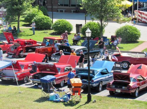 A vibrant car show featuring classic cars with hoods open, people mingling, and a festive outdoor atmosphere.