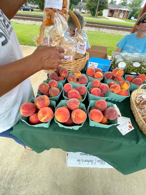 A table at a market displaying baskets of fresh peaches and baked goods, with a person reaching for a bag.