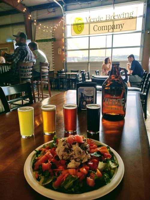 A vibrant salad with tomatoes and chicken sits on a table in a brewery, alongside four colorful beer samples.