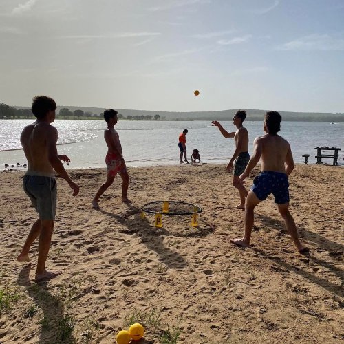 Four shirtless young men play a game of spikeball on a sandy beach near a lake, with a sunny sky in the background.