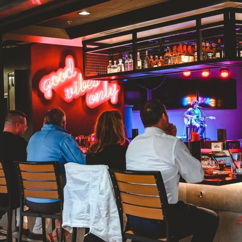 A musician performs on stage in a bar, with a neon sign reading "good vibes only" and an audience enjoying the show.