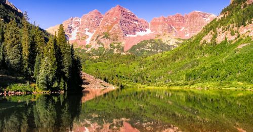 A serene lake reflects majestic mountains and lush greenery under a clear blue sky.