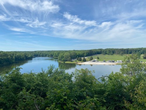 A serene river view surrounded by lush greenery and a sandy beach, with campers visible in the distance under a blue sky.
