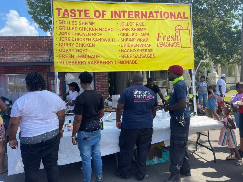 A food stall banner displays various dishes like grilled shrimp, jerk chicken, and fresh lemonade at an outdoor event.