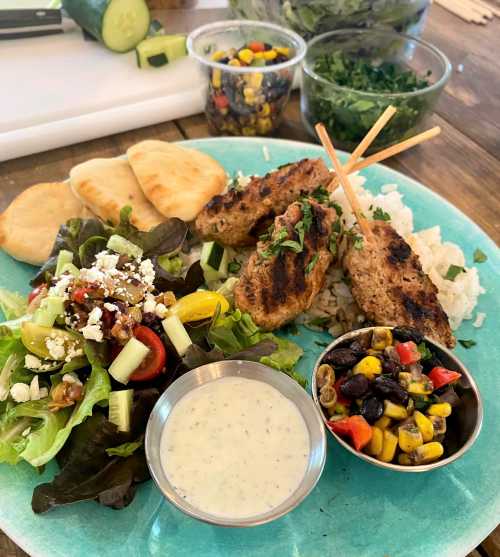 A colorful plate featuring grilled skewers, salad, rice, pita bread, and a side of corn and black bean salad.