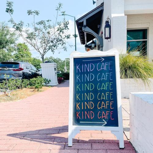 A sidewalk sign outside a café displays "KIND CAFE" in colorful letters, surrounded by greenery and a sunny atmosphere.
