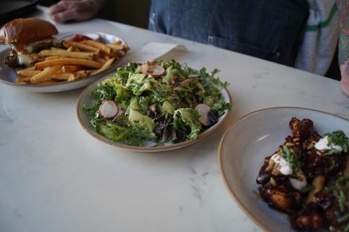 A table with a burger, fries, a fresh salad with radishes, and a plate of sauced chicken wings.