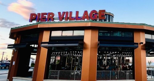 A colorful building with "PIER VILLAGE" signage, featuring a carousel inside and a scenic sky in the background.