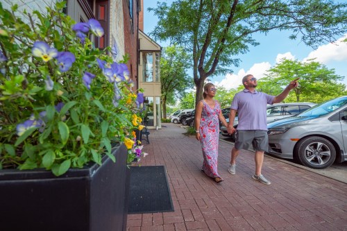 A couple walks hand in hand along a brick path, surrounded by flowers and parked cars under a sunny sky.