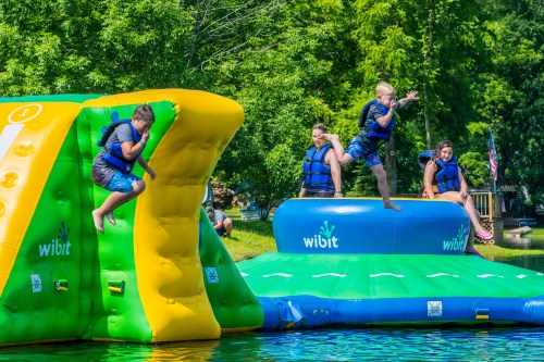 Children playing on inflatable water structures, jumping and climbing in a sunny lakeside setting.