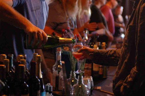 A person pours a drink into a glass at a bar, surrounded by various bottles and people in the background.