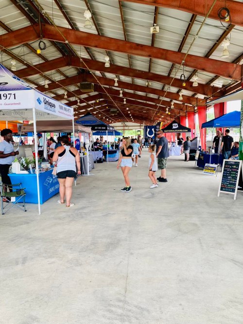 A spacious indoor market with vendors and visitors, featuring booths and colorful banners under a metal roof.