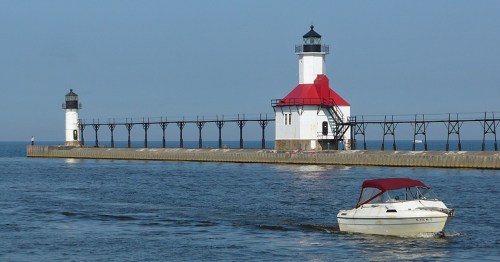 A boat sails past two lighthouses on a pier, with calm waters and a clear blue sky in the background.
